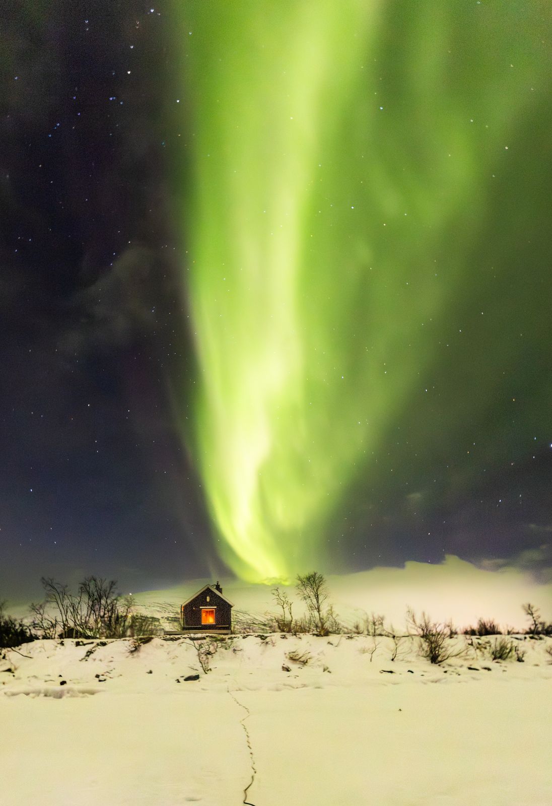 A column of green aurora rising straight up over a lit cabin