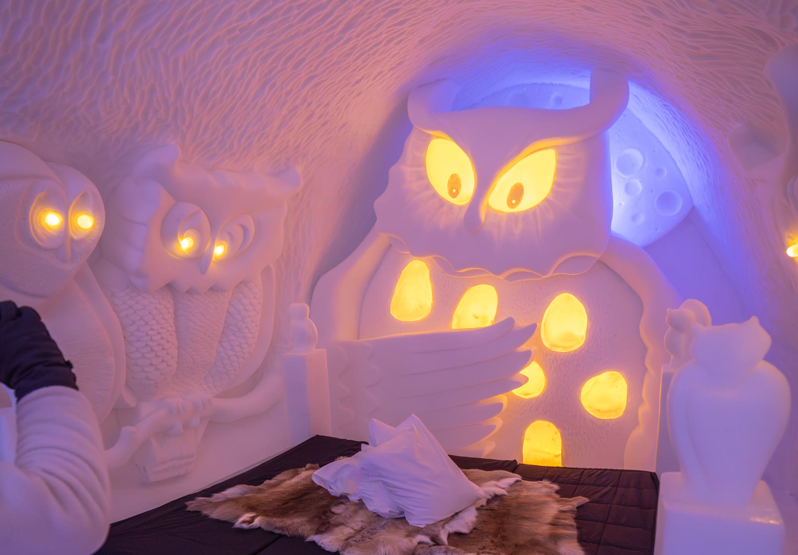 An Icehotel art suite carved from snow and ice, with glowing owls above a bed covered in reindeer hides