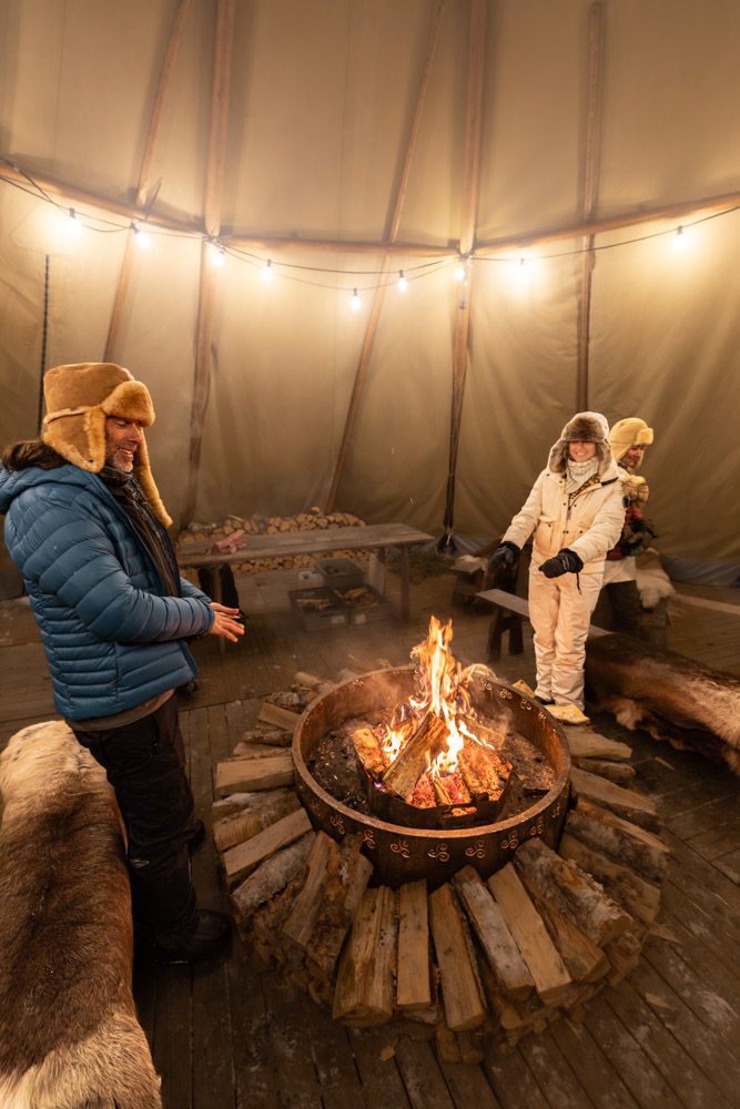 Guests in fur hats around a fire pit inside a lantern-lit tipi