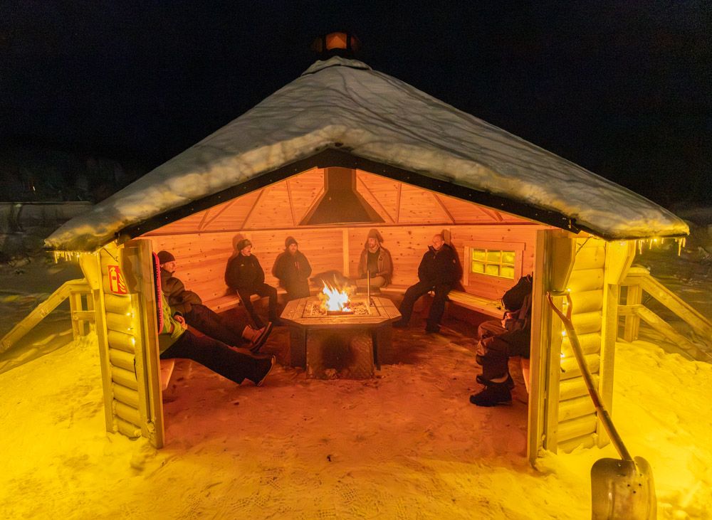 Guests around a fire in a glowing wooden hut at night
