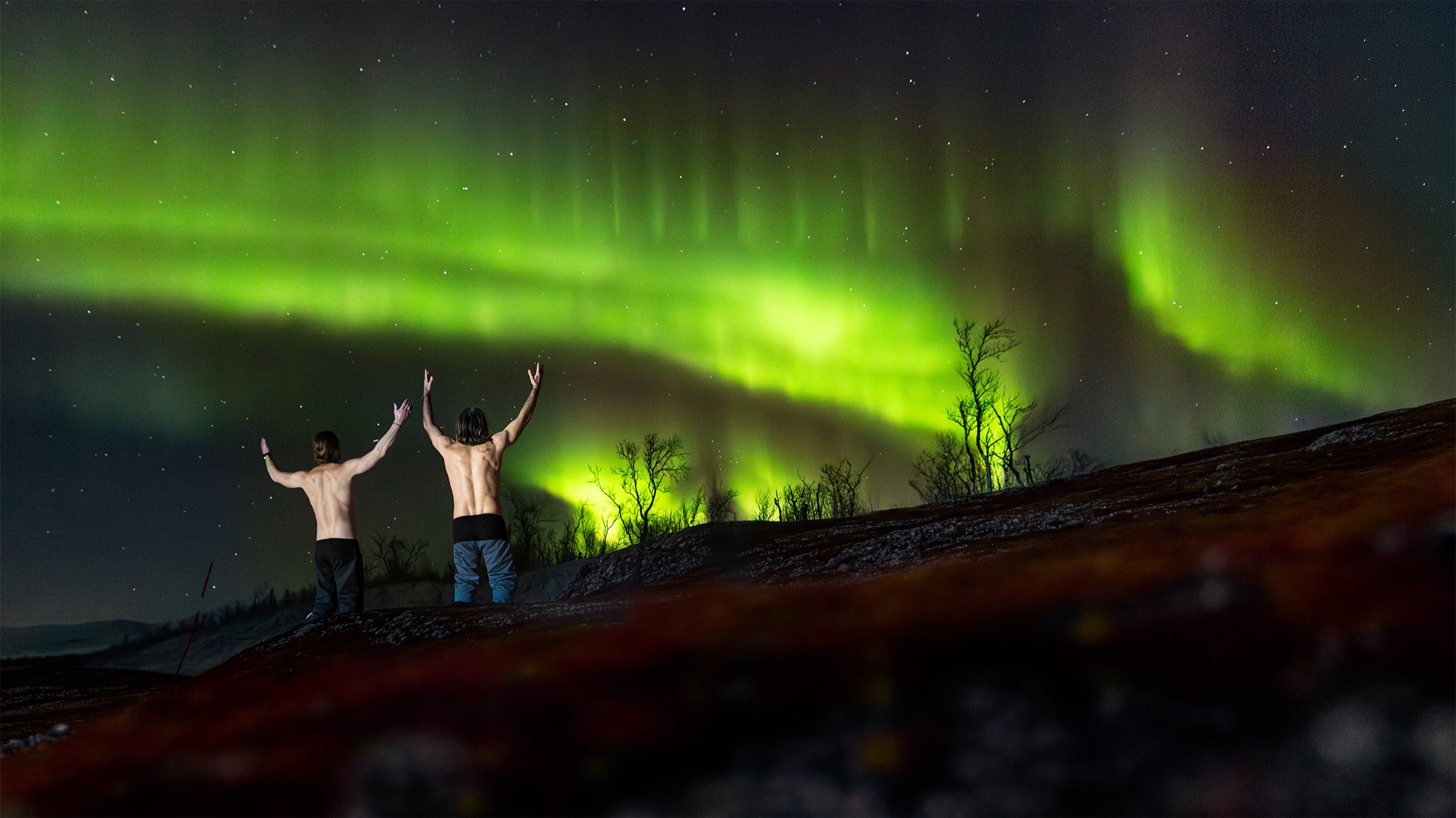 Two Snowcamp guests, shirtless with arms raised, under a bright green aurora
